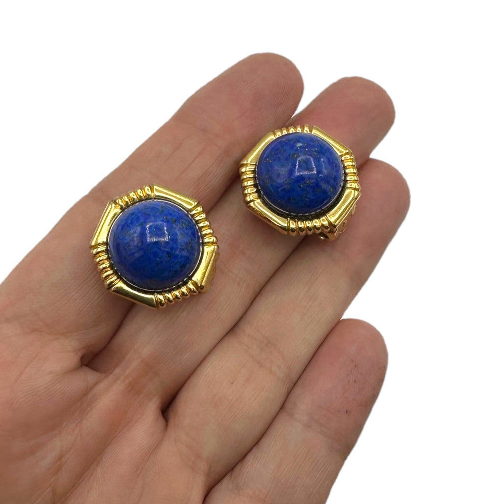 On hand to show size of Vintage Ciner gold clip-on earrings with round blue lapis glass cabochons displayed on white background.