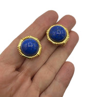 On hand to show size of Vintage Ciner gold clip-on earrings with round blue lapis glass cabochons displayed on white background.