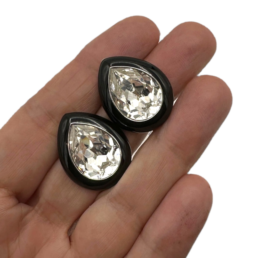 On hand to show size, Vintage Kenneth Jay Lane clear teardrop rhinestone clip-on earrings with black enamel border displayed on white background.