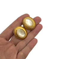 On hand to show size, Monet vintage gold oval clip-on earrings with opalescent cabochon centers and textured gold frame on white background.