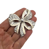 Shown on a hand to show size, Nolan Miller vintage silver bow brooch with white enamel and clear rhinestones, photographed on white background.