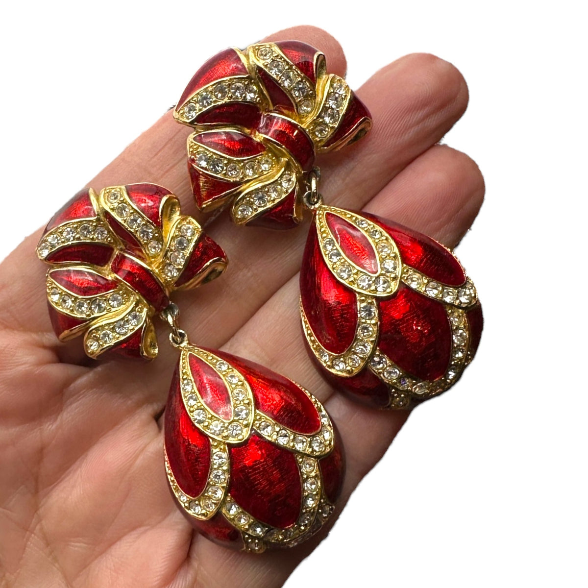 Shown on hand to show size of Whiting & Davis vintage red enamel bow and teardrop rhinestone dangle clip-on earrings, gold-tone statement jewelry on white background.